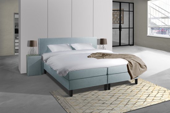 Boxspringbed met ijsblauwe stof, bestaande uit twee boxen, een matras en een hoofdbord. Twee nachtkastjes en een deken liggen erop.
