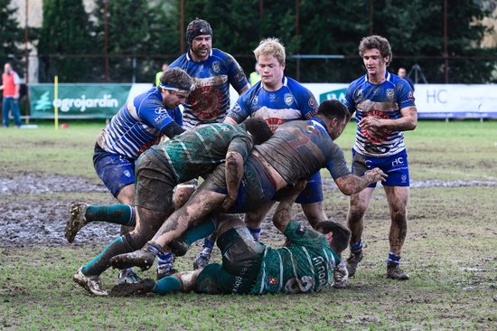 Rugbyspelers in modderige wedstrijd, spelers in blauw-witte en groen-zwarte tenues.