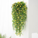 Een weelderige hangende kunstplant met groene bladeren en talloze kleine gele bloemen, hangend tegen een witte muur.