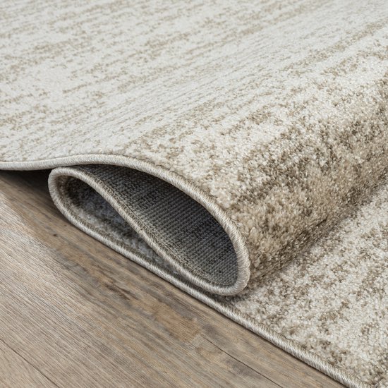 Tapis beige à motif abstrait, enroulé sur un plancher en bois.