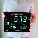 Man in grijs shirt toont een Airvital CO2 monitor met groen display. Toont 579 ppm CO2, 21.6°C en 53.0% luchtvochtigheid.