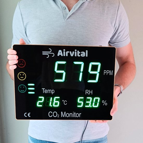 Man in grijs shirt toont een Airvital CO2 monitor met groen display. Toont 579 ppm CO2, 21.6°C en 53.0% luchtvochtigheid.
