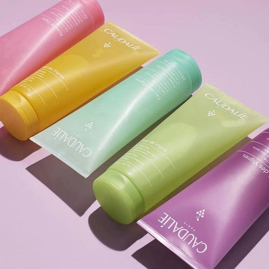 Vijf Caudalie douchegel tubes in roze, geel, mintgroen, lichtgroen en paars, liggend op een lila achtergrond.