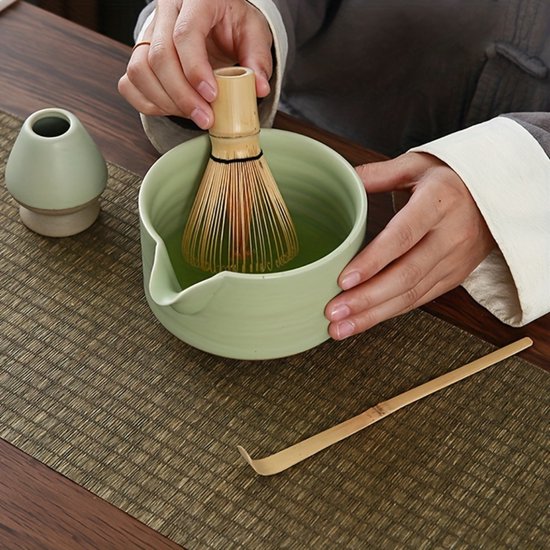 Persoon die matcha klopt in groene kom met bamboe klopper, met kleine blauwe houder.