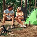 Een stel op zwarte campingstoelen in een bosrijke omgeving bij een tent. Ze kijken elkaar aan.