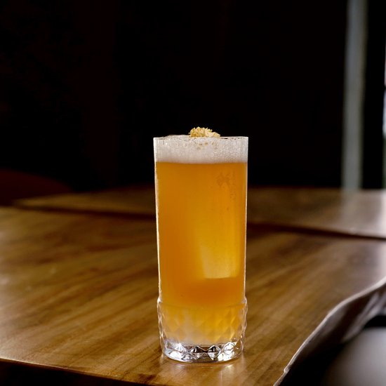 Een hoog, doorzichtig glas met een oranje cocktail en schuim erop, met een diamantvormige bodem.