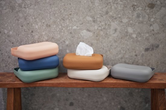 Stapel van verschillende kleuren siliconen doosjes voor billendoekjes op een houten bankje, één doosje met doekjes eruit, een grijs doosje ernaast.