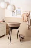 Luma babybadje in beige met zwart onderstel, met een emmer ernaast, in een kinderkamer met commode en kleding aan de muur.