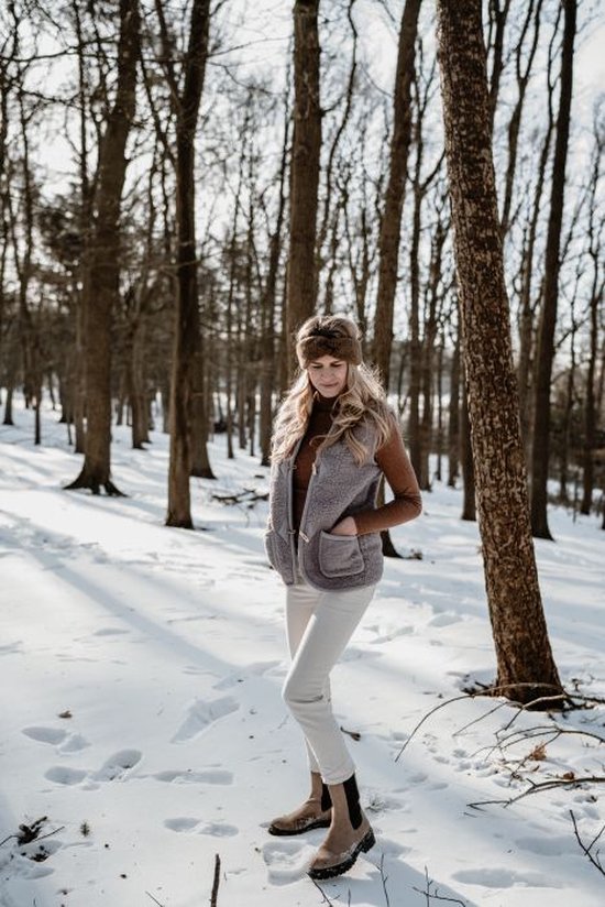 Vrouw in winterlandschap met een lichtgrijze teddy bodywarmer, bruine coltrui en crème broek.