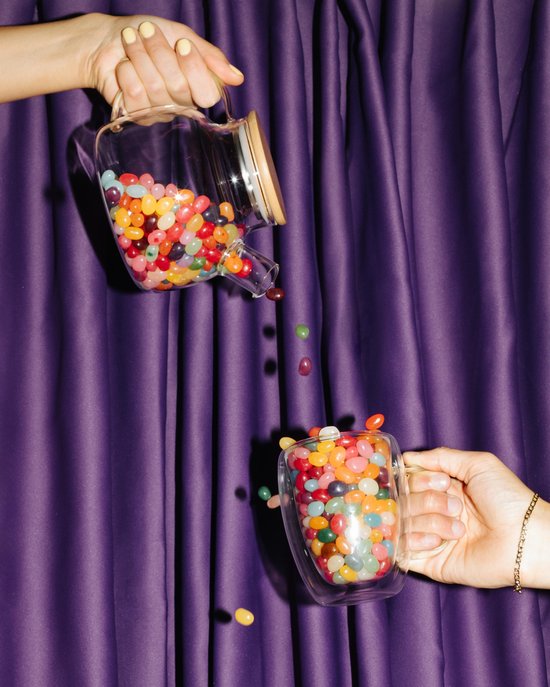 Jelly beans worden uit een glazen schenkkan in een dubbelwandige mok gegoten tegen een paarse achtergrond.