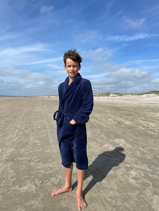Jongen in een donkerblauwe fleeced badjas met ceintuur, staand op het strand.