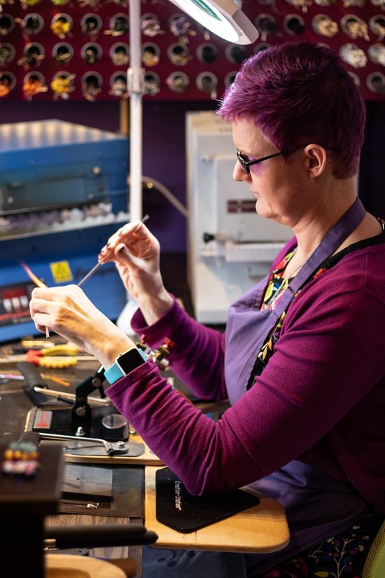 Vrouw met paars haar en een paarse trui, bezig met glasblaaskunst in een werkplaats.