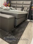 Grijze boxspring met hoog, gestoffeerd hoofdbord, opbergbank en een donkergrijs vloerkleed.