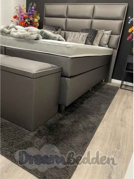 Grijze boxspring met hoog, gestoffeerd hoofdbord, opbergbank en een donkergrijs vloerkleed.