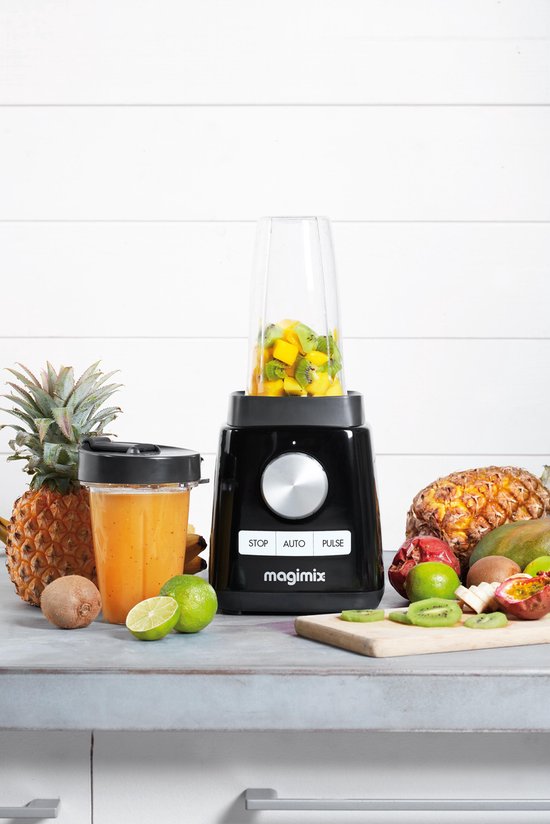 Zwarte Magimix blender met stukjes fruit in de beker, omringd door vers fruit en een glas oranje sap.