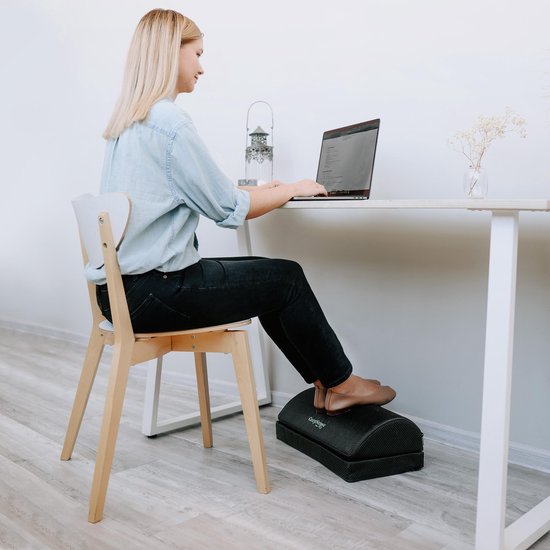 Vrouw met een blauwe blouse en zwarte broek gebruikt een zwarte ergonomische voetensteun onder haar bureau, terwijl ze op een laptop typt.