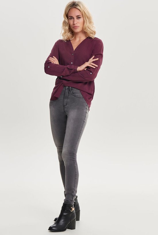 Vrouw met armen gekruist in grijze skinny jeans en bordeauxrode blouse.