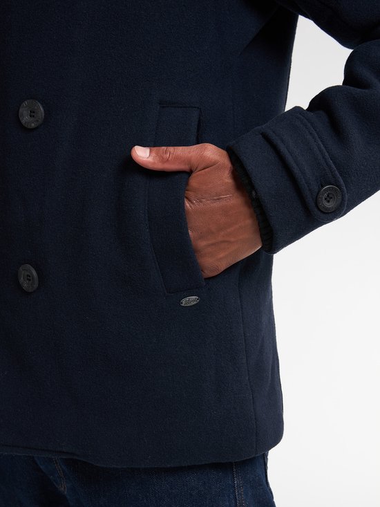 Detail van de zak van een donkerblauwe peacoat, met een hand erin.