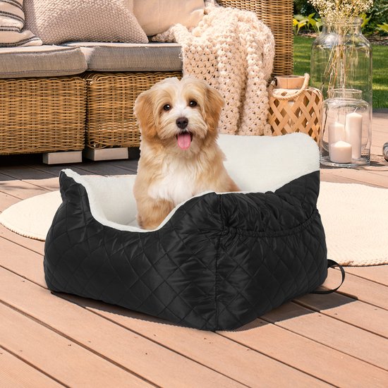 Een beige hondje zit in een zwart, gewatteerd autostoeltje voor huisdieren met witte pluche voering. Het stoeltje staat op een houten terras.