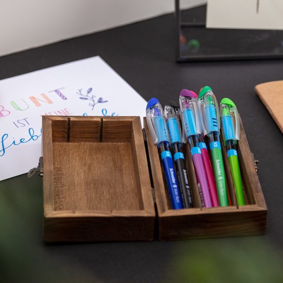 Blauwe, roze, groene en zwarte Schneider Slider Basic pennen in een houten doosje naast een brief met de tekst 'BUNT IST MEINE LIEBLINGSFARBE'.