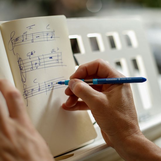Een hand schrijft muziek op een notitieboek met een blauwe Schneider Slider Basic balpen.
