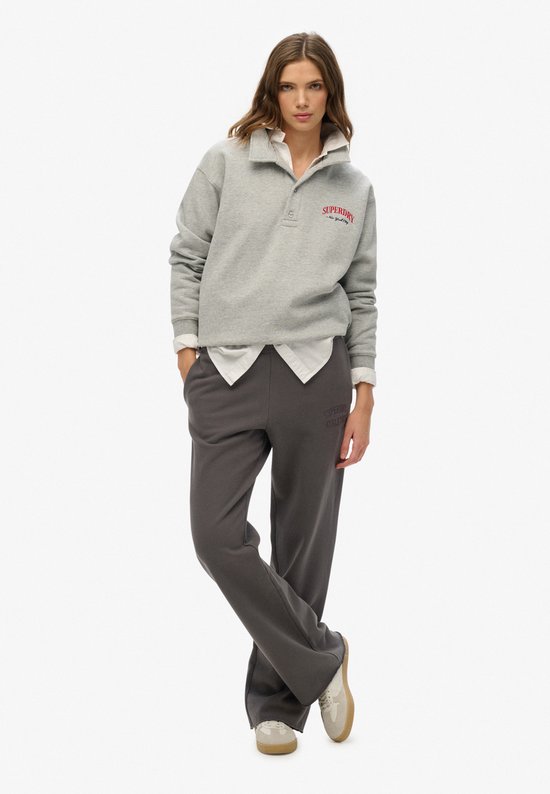 Model draagt een grijze Superdry sweater en bijpassende grijze joggingbroek met wijde pijpen en geborduurd logo.