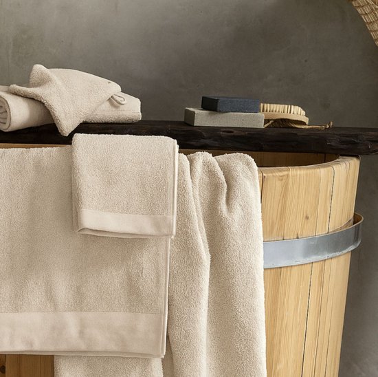 Verschillende beige handdoeken, waaronder opgerolde handdoeken en een handdoek die over een houten ton hangt. Ook zeep en een borstel zijn zichtbaar.