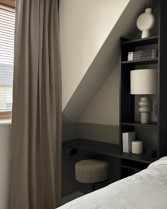 Taupe gordijn hangt voor een raam naast een bureau met een bijzettafel en boekenplank in een slaapkamer.
