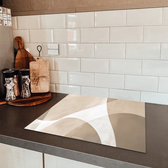 Abstracte beige beschermhoes voor inductiekookplaat op een donker aanrecht, met witte metrotegels op de achtergrond.