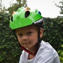 Lichtgroene kinderfietshelm met dinosaurusprint, grote blauwe ogen en scherpe tanden. Een jongen draagt de helm.