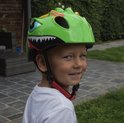 Lichtgroene kinderfietshelm met dinosaurusprint, grote blauwe ogen en scherpe tanden. Een jongen draagt de helm.
