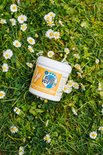 Potje Blije Voeten eelt- en klovencrème in gras met madeliefjes. Het label toont een blauwe voetprint met de tekst "Blije Voeten" en een afbeelding van voeten.