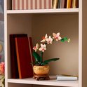 LEGO orchidee in een pot op een boekenplank, naast boeken en een vaas met bloemen.