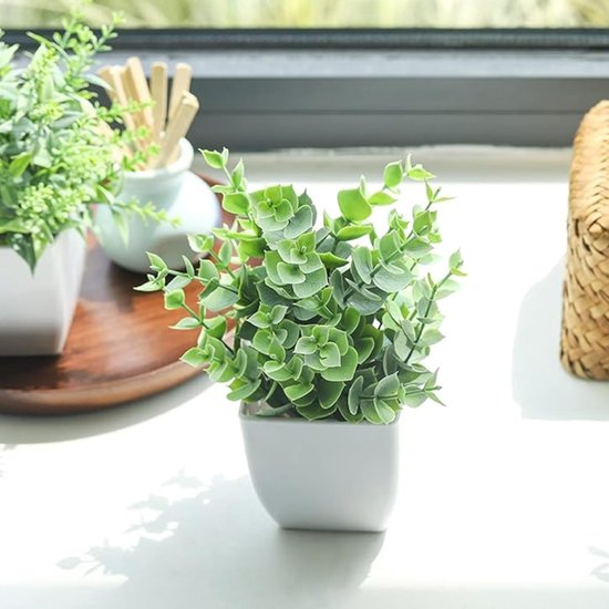 Een kunstplant met dicht op elkaar staande, grote ronde groene bladeren in een witte vierkante pot. Staat op een vensterbank naast een rieten mand.