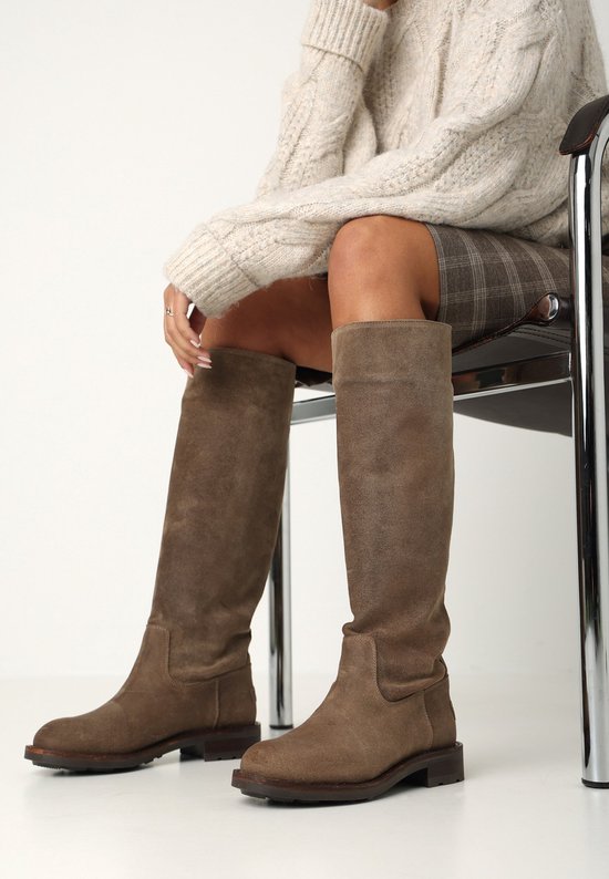Model zittend op een stoel, draagt taupe suède kniehoge laarzen, een geruite rok en een gebreide trui.