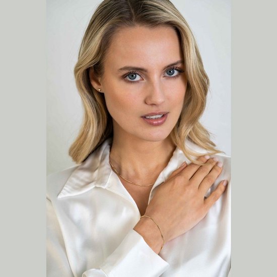 Model met blonde haren draagt een gouden ketting en armband, witte blouse.