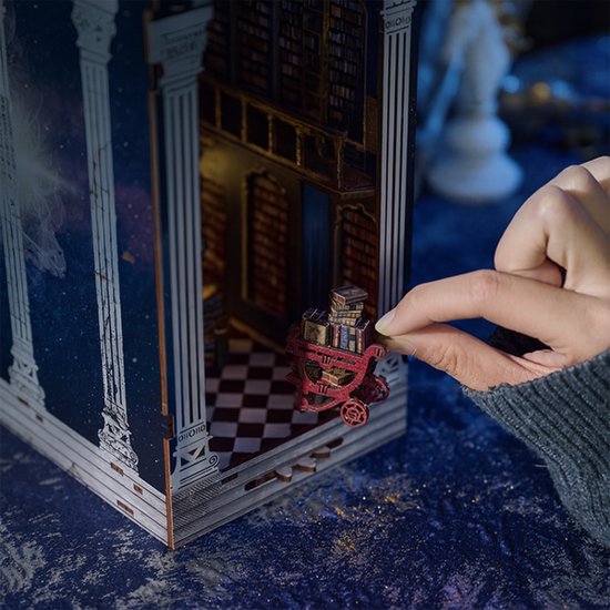 Hand plaatst miniatuur boekentrolley in een model van een bibliotheek met boekenkasten.