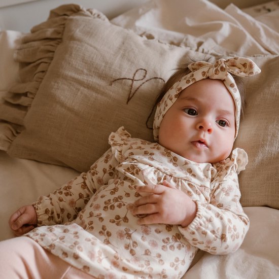 Baby ligt op bed met een beige romper met bloemenprint en bijpassende hoofdband.