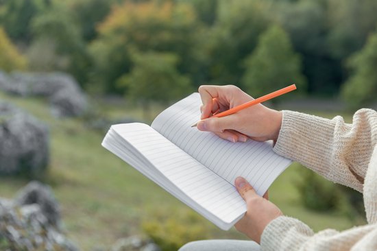 Personne écrivant dans un carnet ouvert ligné, à l'extérieur dans un environnement naturel.