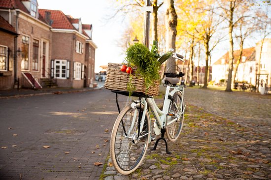 Lichtgroene fiets met rieten mand met groente en twee rode tomaten op een geplaveide straat.