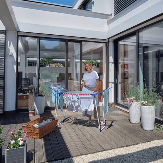 Vrouw op een houten terras plaatst kleding op een wit droogrek met blauwe en rode accenten.