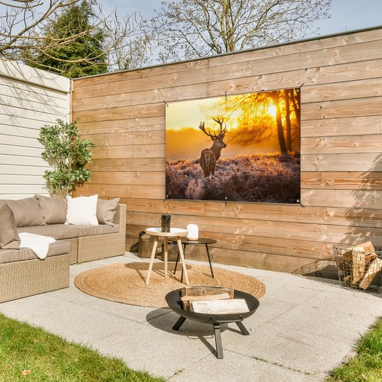 Een tuinposter met een hert in de ochtendzon hangt aan een houten schutting boven een lounge set.