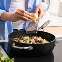 Vrouw gratineert gehaktballetjes, pasta en broccoli in een zwarte hapjespan met Parmezaanse kaas.