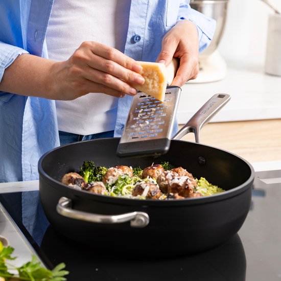 Vrouw gratineert gehaktballetjes, pasta en broccoli in een zwarte hapjespan met Parmezaanse kaas.