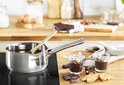 Roestvrijstalen steelpan met een houten lepel en chocolade, naast twee glaasjes.