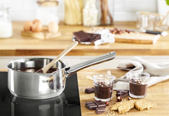 Roestvrijstalen steelpan met een houten lepel en chocolade, naast twee glaasjes.