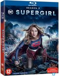 Blu-ray hoes van Supergirl Seizoen 3 met de actrice in haar Supergirl-kostuum, dat een rood met blauw shirt met het 'S'-logo, een rode cape, een rode rok en kniebeschermers bevat. Ze knielt op een stadsruïne in een dynamische pose, terwijl explosies op de achtergrond te zien zijn. De tekst 'Supergirl Season 3' staat centraal.