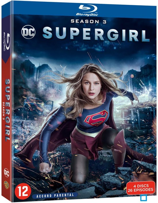 Blu-ray hoes van Supergirl Seizoen 3 met de actrice in haar Supergirl-kostuum, dat een rood met blauw shirt met het 'S'-logo, een rode cape, een rode rok en kniebeschermers bevat. Ze knielt op een stadsruïne in een dynamische pose, terwijl explosies op de achtergrond te zien zijn. De tekst 'Supergirl Season 3' staat centraal.
