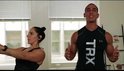 Vrouw met donker haar die de TRX-banden gebruikt, terwijl een man ernaast staat te praten.
