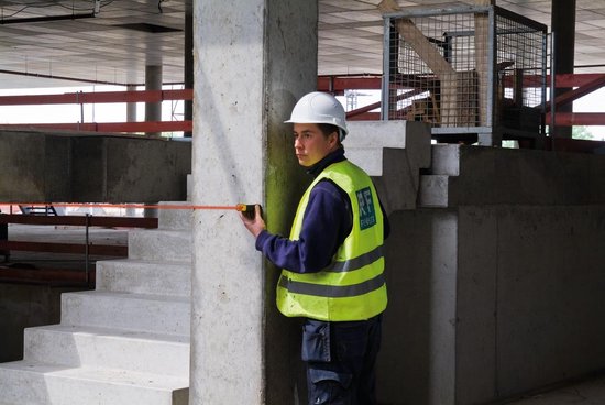 Bouwvakker in gele veiligheidshesje meet met een rolmaat langs een betonnen pilaar.